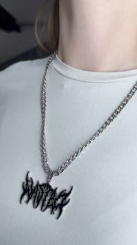 Srebrna lančić sa crnim privjeskom u obliku plamena koji sadrži stilizirani tekst. Pozadina je svijetla košulja.