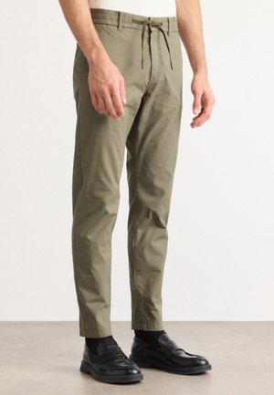 Mann trägt olivgrüne Kordelzughose und schwarze Loafers, steht auf einem hellgrauen Boden vor einem schlichten Hintergrund.