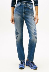 Mujer usando jeans azules con bordado floral en los muslos y zapatillas deportivas moradas, de pie contra un fondo blanco liso.