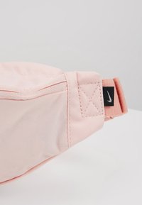 Ledvinka Nike v světle růžové tkanině s hladkým povrchem. Obsahuje šité zapínání na zip a černou štítkovou značku na popruhu.