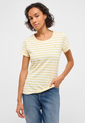 Frau mit kurzen lockigen Haaren, die ein weißes und blassgelb gestreiftes T-Shirt und blaue Jeans trägt, mit einer Hand in der Hosentasche.