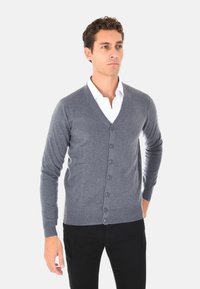Cardigan grigio in lana con design a V, sei bottoni frontali e maniche lunghe. Indossato sopra una camicia bianca, abbinato a pantaloni neri.