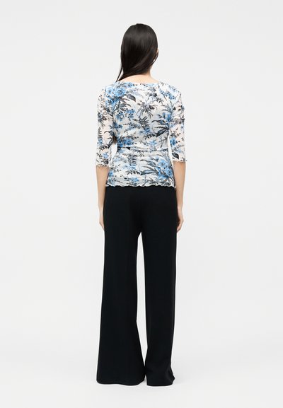 Γυναίκα με μακριά σκούρα μαλλιά, που φοράει λευκή και μπλε φλοράλ μπλούζα με μανίκια τριών τετάρτων και μαύρο παντελόνι φαρδιάς γραμμής, απεικονίζεται από πίσω.