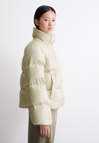 Puffjakke i myk beige, med høy krage, kviltet mønster og sidelommer. Materialet ser blankt og isolert ut.