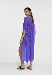 Kaftan viola, leggero, con maniche a pipistrello, vita arricciata e fessure laterali. Il modello indossa sandali neri e grandi orecchini rosa.