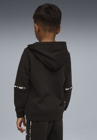 Niño con el pelo corto, llevando una sudadera negra con capucha y pantalones a juego con logotipos blancos, de espaldas frente a un fondo gris.