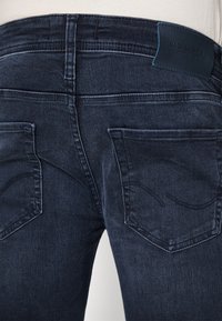 Jeans de mezclilla azul oscuro con una textura suave, dos bolsillos traseros y una etiqueta de logo en la cintura. El diseño tiene detalles de desvanecimiento sutiles.