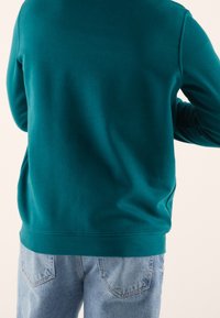 Sininen vihertävän sävyinen pusero, jossa pitkät hihat, resorit hihansuuissa ja helmassa. Yhdistetty vaaleansinisiin farkkuihin, joissa on takataskut ja kulutettu pinta.