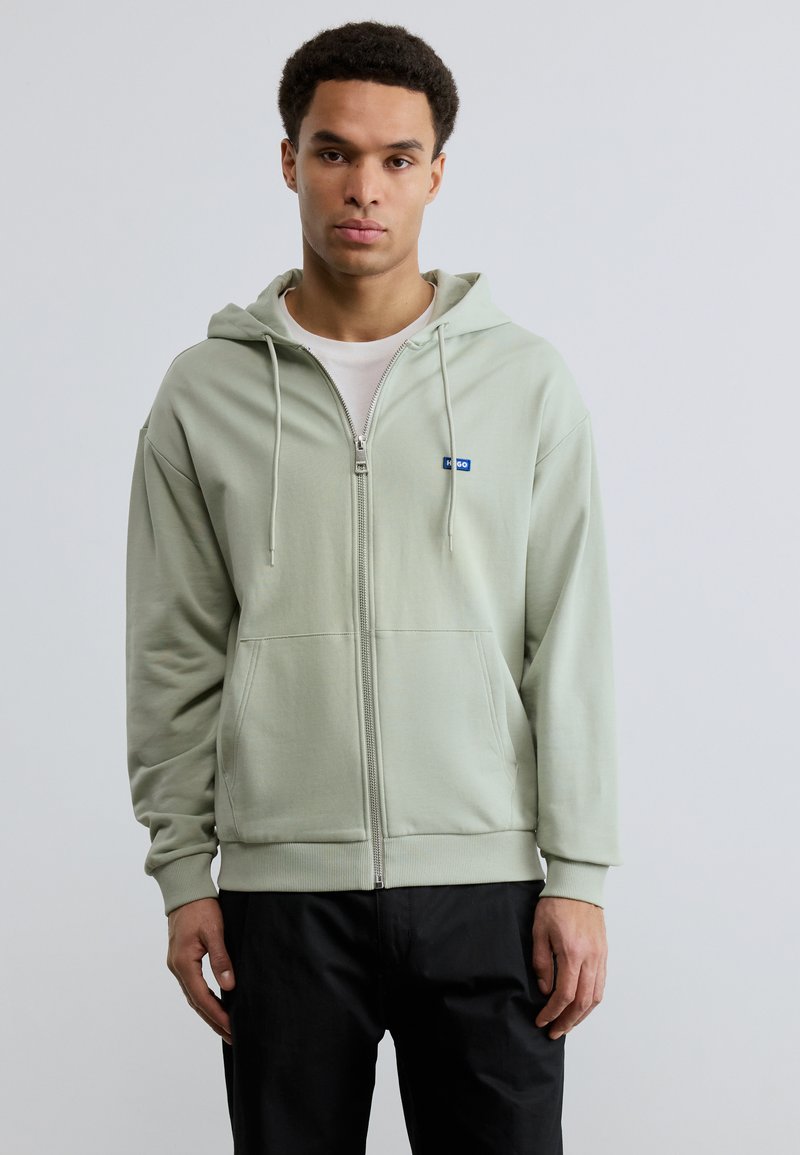 Νεαρός άνδρας που φοράει ένα ανοιχτό πράσινο φούτερ με φερμουάρ πάνω από λευκό πουκάμισο και μαύρο παντελόνι, στέκεται μπροστά σε ένα απλό φωτεινό φόντο.