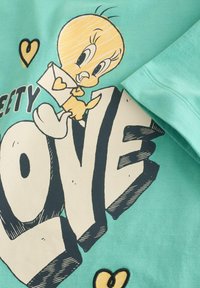 Hellgrünes T-Shirt mit einem cartoonhaften gelben Vogel, der einen Umschlag hält, mit dem Wort "LIEBE" in fetten schwarzen Buchstaben und kleinen gelben Herzen.