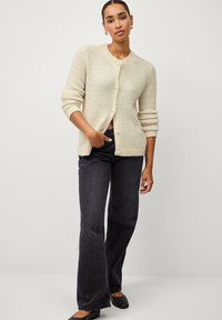 Cardigan en tricot beige à col rond, boutons en perles et poignets côtelés, associé à un jean en denim foncé et des ballerines noires.
