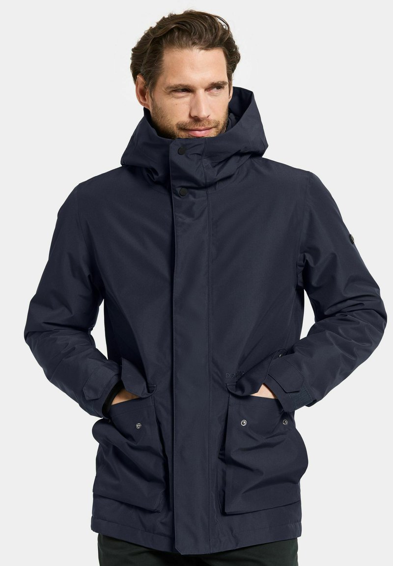 Veste imperméable bleu marine avec un col haut, fermeture à pression à l'avant et deux grandes poches latérales. Texture lisse avec des poignets ajustables.