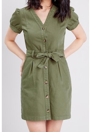 Femme portant une robe olive à boutons sur le devant, avec des manches bouffantes et une ceinture nouée à la taille, debout les mains sur les hanches.