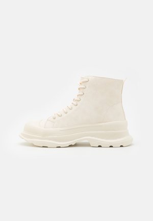 Beżowy wysoki but typu sneaker z grubą, teksturowaną podeszwą i sznurowaniem z przodu, widoczny z boku na jednolitym jasnym tle.