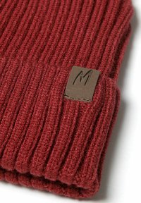 Berretto a coste rosso con un'etichetta in pelle marrone che presenta un "M." in rilievo. La trama del tessuto è morbida e ben intrecciata.