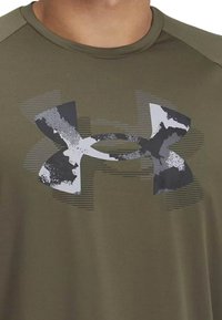 Camiseta deportiva de color verde oliva con mangas cortas, que presenta un gran logo texturizado en blanco y negro en el centro, y sutiles rayas horizontales en el fondo.