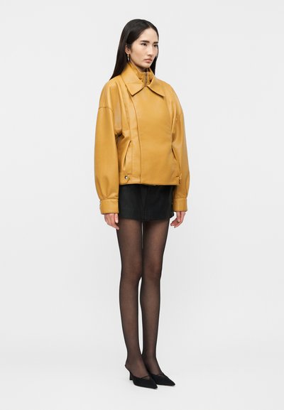Γυναίκα όρθια που φοράει ένα υπερμεγέθες δερμάτινο μπουφάν μουσταρδί, μαύρη μίνι φούστα, διάφανα μαύρα καλσόν και μαύρα μυτερά παπούτσια.