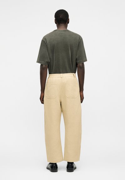 Homme portant un pantalon beige ample avec poches arrière, une chemise à manches courtes vert olive foncé et des chaussures de ville noires, debout devant un fond blanc.