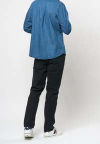 Camisa de denim en azul, de mangas largas, con dobladillo curvado. Pantalones negros oscuros, ajustados. Zapatillas blancas con acentos en azul y rojo, suela plana.