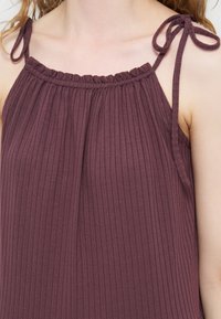 Κοντινή λήψη ενός μωβ ριμπ πλεκτού αμάνικου τοπ με σούρες στο ντεκολτέ και δεμένες τιράντες στους ώμους σε άτομο με ανοιχτόχρωμο δέρμα.