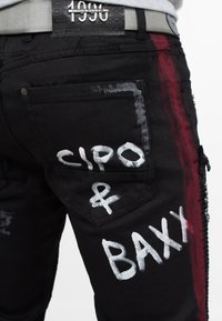 Jeans in denim nero con testo in stile graffiti bianco "CIPO & BAXX", accenti di vernice rossa e dettagli testurizzati sulle tasche. Presenta una cintura argentata.