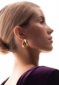 Orecchino a cerchio d'oro con un design liscio e ondulato. Modella con capelli castano chiaro, indossando un top viola scuro, in profilo su uno sfondo bianco.