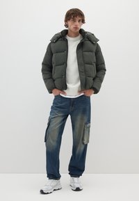 Giacca a vento di colore verde scuro con chiusura a zip e cappuccio, abbinata a pantaloni cargo in denim blu con tasche laterali e vestibilità comoda. Sneakers bianche.