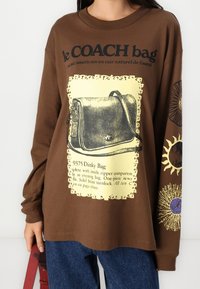 Ruskea pitkähihainen t-paita, jossa on mustan käsilaukun kuvasto keltaisella tekstillä, koristeellisilla reunoilla ja värikkäillä aksenttikuvioilla hihoissa.