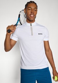 Biele výkonnostné polo s golierom na polovičný zips, obsahuje textúrovaný vzor, logo na hrudi, nosené s modrými atletickými šortkami, drží tenisovú raketu.