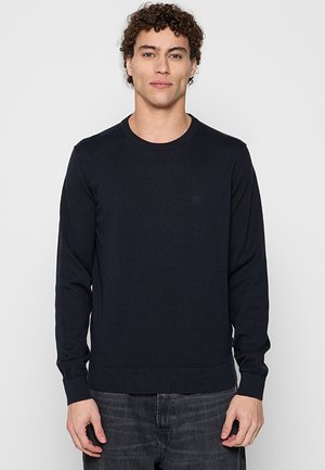 Junger Mann mit lockigem Haar trägt einen schlichten schwarzen Langarmpullover und dunkle Jeans, steht vor einem hellgrauen Hintergrund.