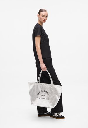 Borsa a tracolla argento con testo stampato nero, dotata di due manici. Il modello indossa una maglietta nera e pantaloni, con sneaker in bianco e nero.
