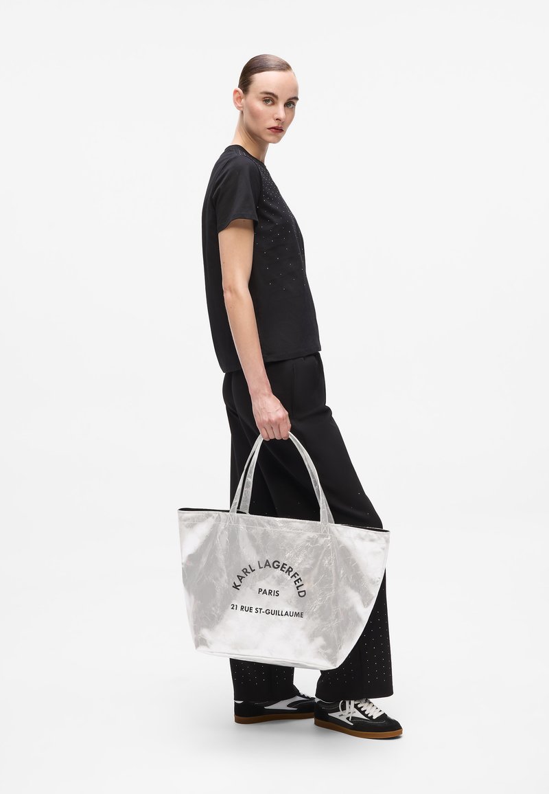 Sac fourre-tout argenté avec texte imprimé noir, doté de deux poignées. Le modèle porte une chemise noire et un pantalon noir, avec des baskets noir et blanc.