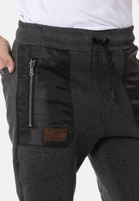 Dark gray jogger pants with a textured fabric, featuring a black smooth pocket with a metal zipper and a brown leather logo patch.