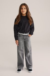 Svart långärmad skjorta med texturerad detalj, grå vida jeans med lätt tvätt, vit bälte och svarta sneakers med vita sulor.