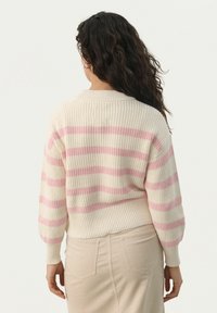 Mäntel mit grobem Strick in Creme mit pinken Streifen, rundem Ausschnitt und gerippten Bündchen. Kombiniert mit einem beigen Rock.