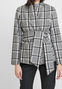Blazer a scacchi grigio con dettagli neri e arancioni, caratterizzato da un design avvolgente e una cintura in vita. Indossato sopra un top grigio e pantaloni a righe.