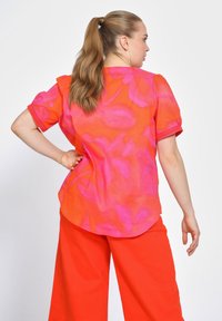 Korte mouwen, losse shirt in fel oranje met een roze bloemenpatroon; gecombineerd met wijde, rode broeken. Glad fabric, casual ontwerp.