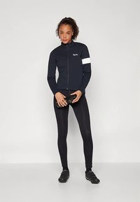Veste de sport bleu marine avec fermeture éclair, col montant et accent blanc sur la manche ; associée à un legging noir et des chaussures de sport ; tenant une bouteille d'eau noire.