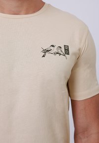 Μπεζ βαμβακερή μπλούζα με μαύρη εικονογράφηση δύο πουλιών που κάθονται σε ένα κλαδί, με ιαπωνικό κείμενο και μια μικρή λουλουδάτη λεπτομέρεια.