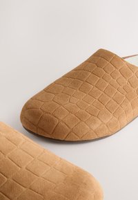 Gros plan sur des chaussures slip-on en daim beige avec un motif texturé à écailles de crocodile et des bouts carrés arrondis sur une surface unie.