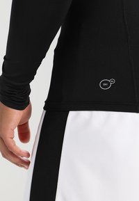 Sort langærmet skjorte lavet af fugtabsorberende stof, med et synligt "DRY CELL" logo, parret med hvide shorts med sorte accenter.