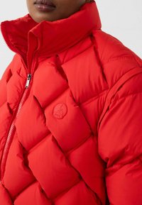 Rote Steppjacke mit hohem Kragen, gesteppten Abschnitten, Reißverschluss und einem Logobadge auf der linken Seite. Glatte Textur.