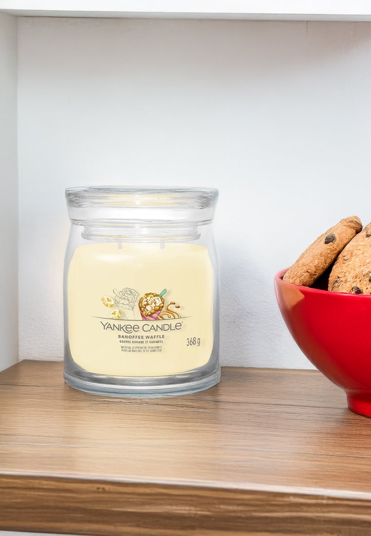 Candelina Yankee in un barattolo di vetro trasparente, etichettato "Banoffee Waffle", con cera di colore crema; accompagnata da una ciotola rossa di biscotti con gocce di cioccolato.