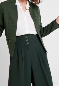 Femme portant un blazer texturé vert foncé, un pantalon taille haute vert foncé avec quatre boutons, et une chemise blanche boutonnée sur un fond clair.