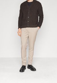 Brun texturerad cardigan med V-ringning och svarta knappar, parat med beigefärgade byxor och svarta loafers. Minimalistisk design, figurnära silhuett.