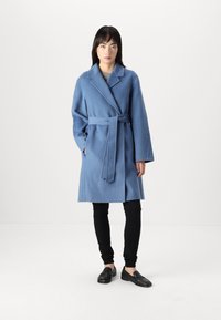 Abrigo de mezcla de lana azul claro con cintura ceñida por un cinturón, cuello oversize y mangas largas, combinado con pantalones negros ajustados y mocasines negro.