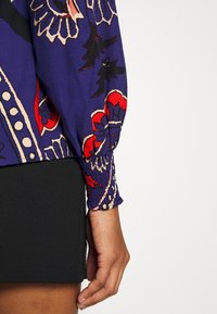 Paarse gebloemde blouse met smock manchetten, voorzien van bloemenpatronen in rood, zwart en beige; gecombineerd met een zwarte rok.