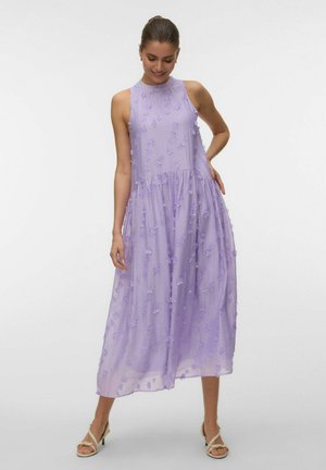Femme portant une robe midi lilas sans manches avec des détails floraux texturés, debout avec une main sur la hanche et regardant vers le bas en souriant.