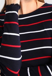 Mörkblå tröja med röda och vita horisontella ränder, med båt-utsnitt och volangärmar. Tillverkad av ett mjukt, stretchigt material.