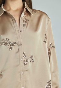 Blusa di satin in beige chiaro con colletto, caratterizzata da ricami floreali in paillettes nei toni dell'oro e del marrone, chiusa con bottoni sul davanti.